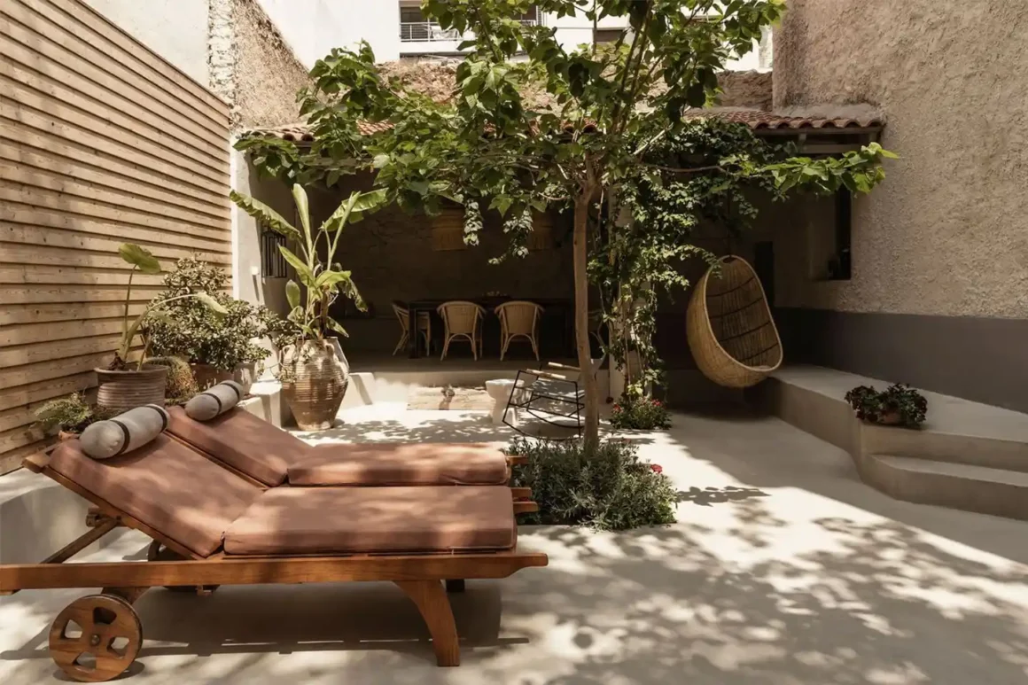 Cortile urbano in stile mediterraneo con lettini in legno, albero da ombra, pavimento in resina e zona pranzo coperta da rampicanti